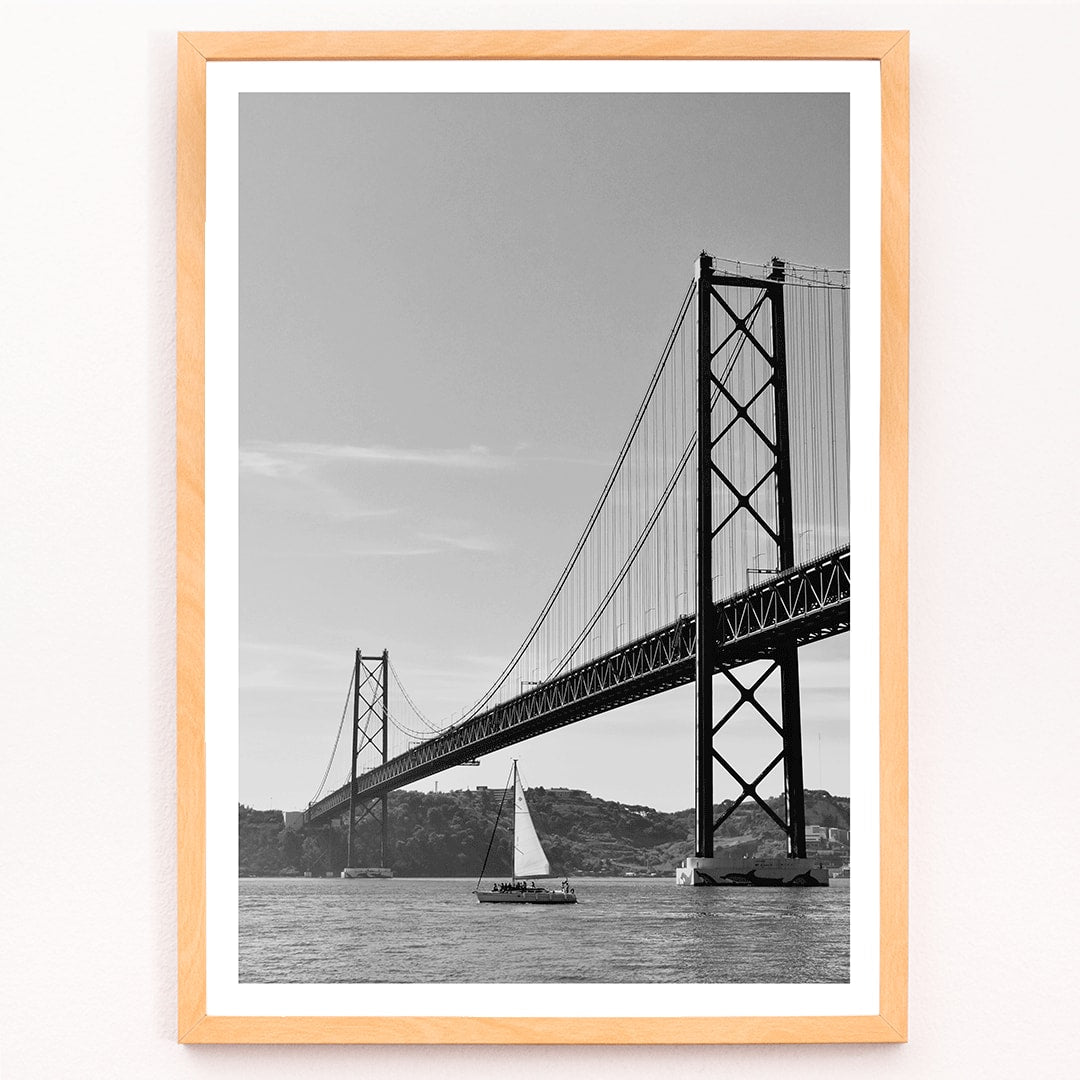 Lisbon Bridge Black and White Poster by Raphael Alves featuring a framed photograph of a suspension bridge and sailboat in the water, framed and displayed in modern living room decor