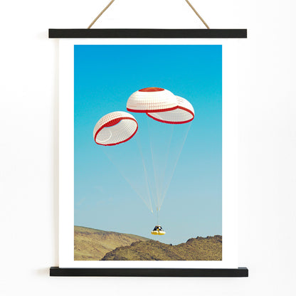 CST-100 crew capsule landing with three parachutes over desert landscape by NASA and The Boeing Company poster artwork, with wooden magnetic frame on white background